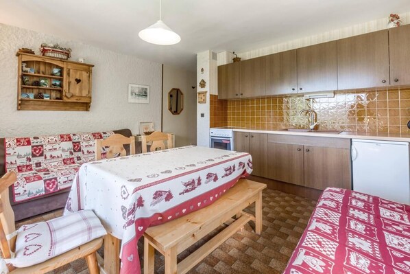 A cozy room with a wooden bench, a table with a patterned tablecloth, a kitchenette with wooden cabinets, and a tiled backsplash.