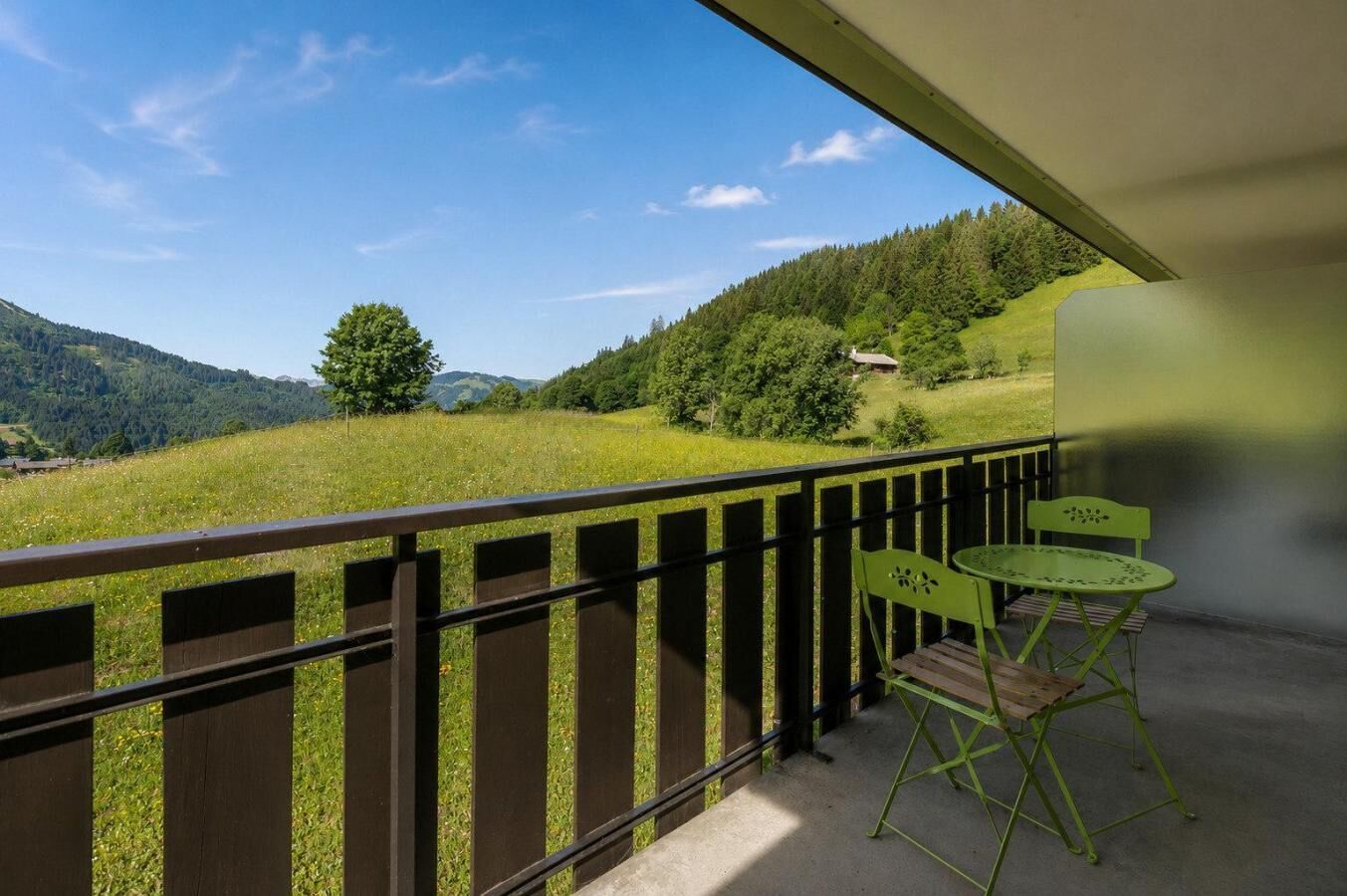 A balcony with a view of a green landscape, a wooden railing, and a table with two chairs.