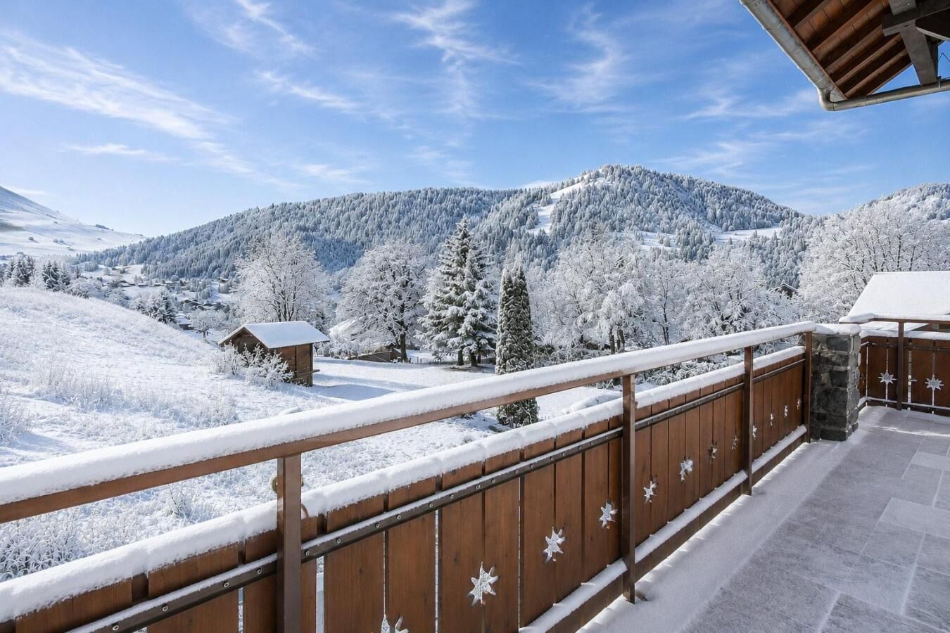 A wooden balcony with snow-covered ground, a stone wall, and a view of snow-laden trees and mountains.