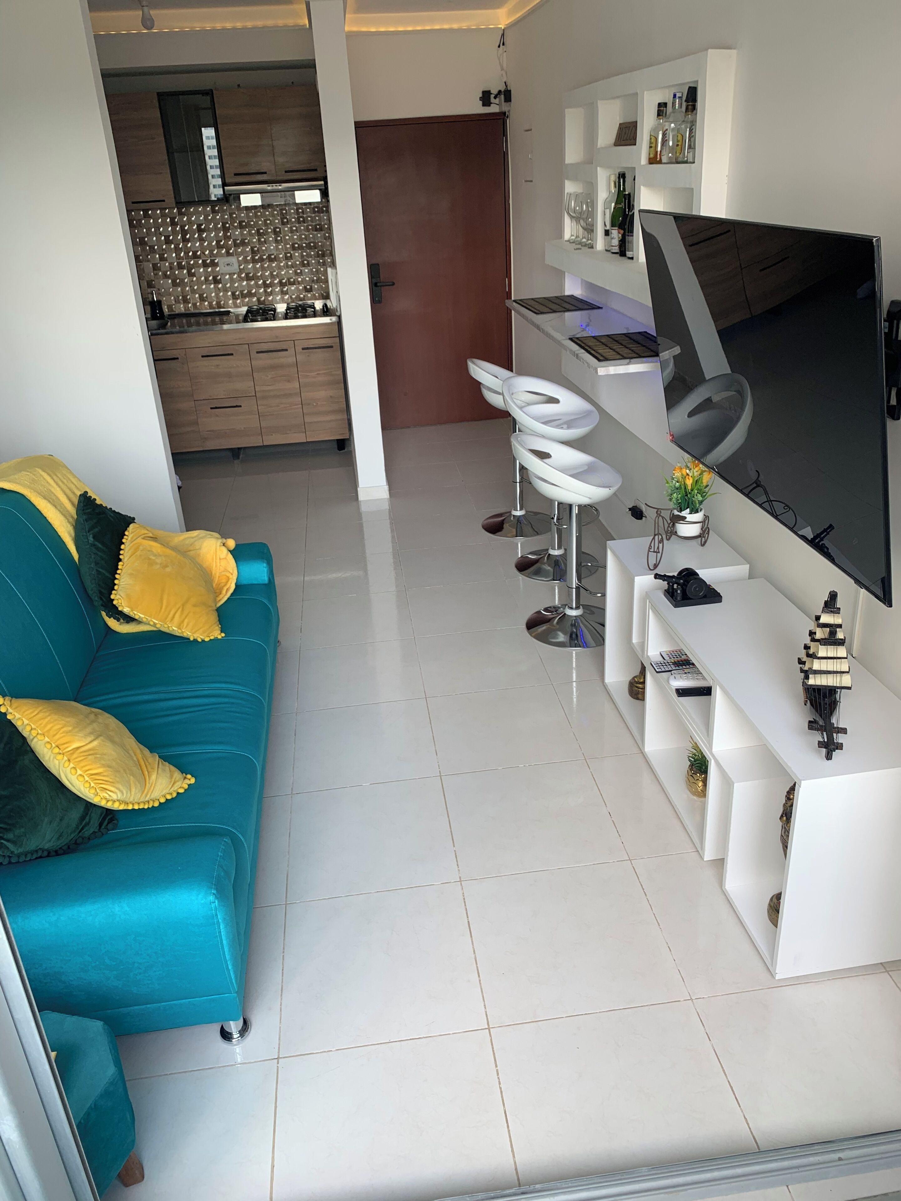 A modern living room with a blue sofa, white bar stools, a large flat-screen TV, and a kitchen in the background.
