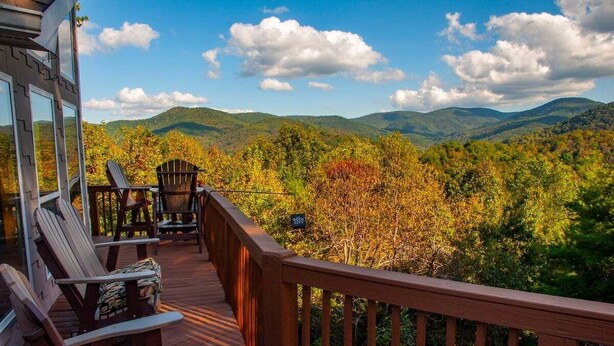 A wooden deck with Adirondack chairs and a table, overlooking a forested mountain range.