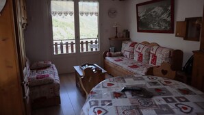 A cozy room with a bed, a sofa, and a small table. There are two windows with curtains, a painting on the wall, and a wooden door. - 2nd floor, balcony, parking, tv, ski locker, 40m², Valloire (Valloire)