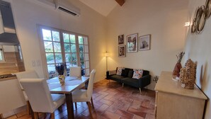 Ein Essbereich mit Tisch und Stühlen, einer Couch und einer Lampe. Im Hintergrund sind Fenster und eine Küche zu sehen.