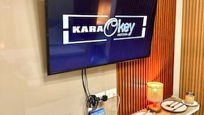 Ein Zimmer mit montiertem Fernseher, der 'KARA Okkey' zeigt, und einer Spiele-Einrichtung inklusive PlayStation 4 Konsole, Controllern und einem Spielgehäuse.