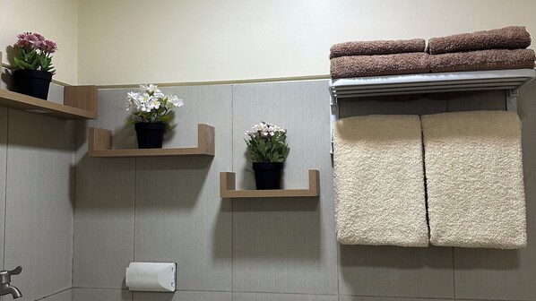 Une salle de bain avec deux étagères en bois où sont posées des plantes en pot, un porte-serviettes avec des serviettes pliées et un support à papier toilette.