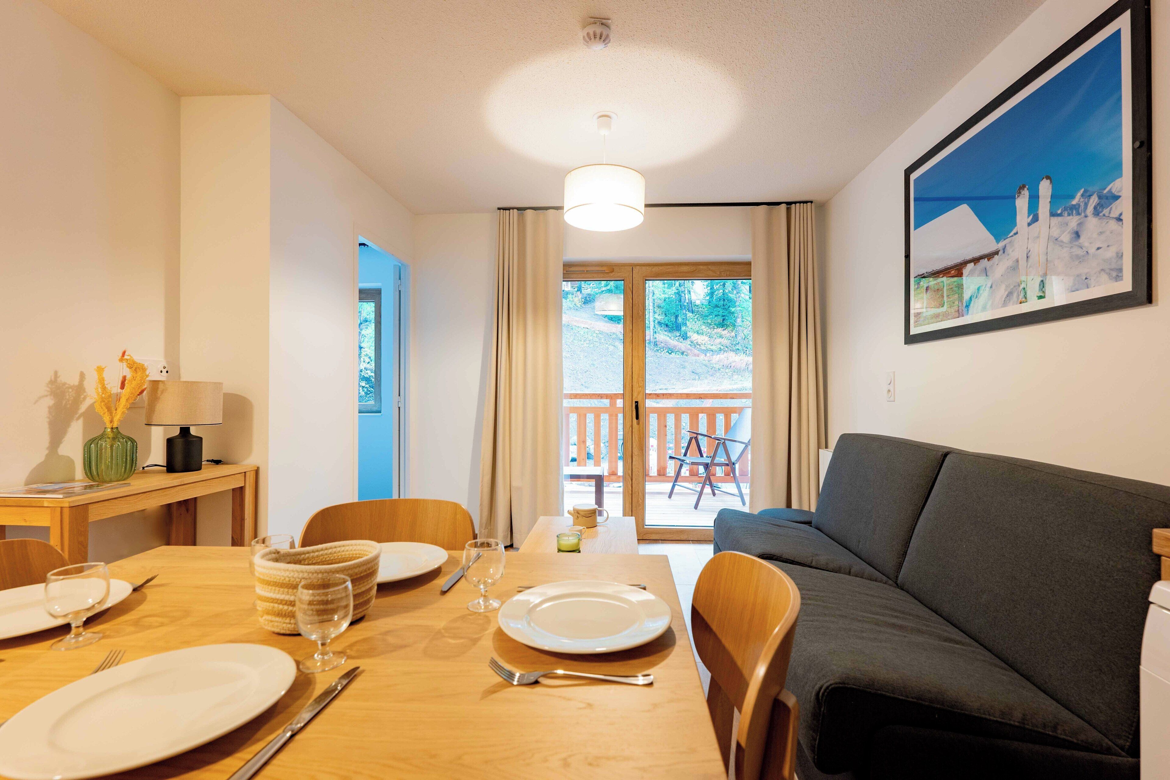 A dining area with a wooden table set for four, a sofa, and a painting of a snowy mountain scene.