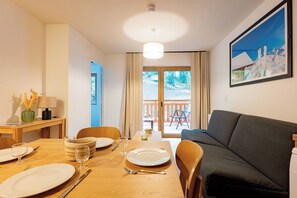 A dining area with a wooden table set for four, a sofa, and a painting of a snowy mountain scene. - Résidence Arya - Living Stone (Vars)