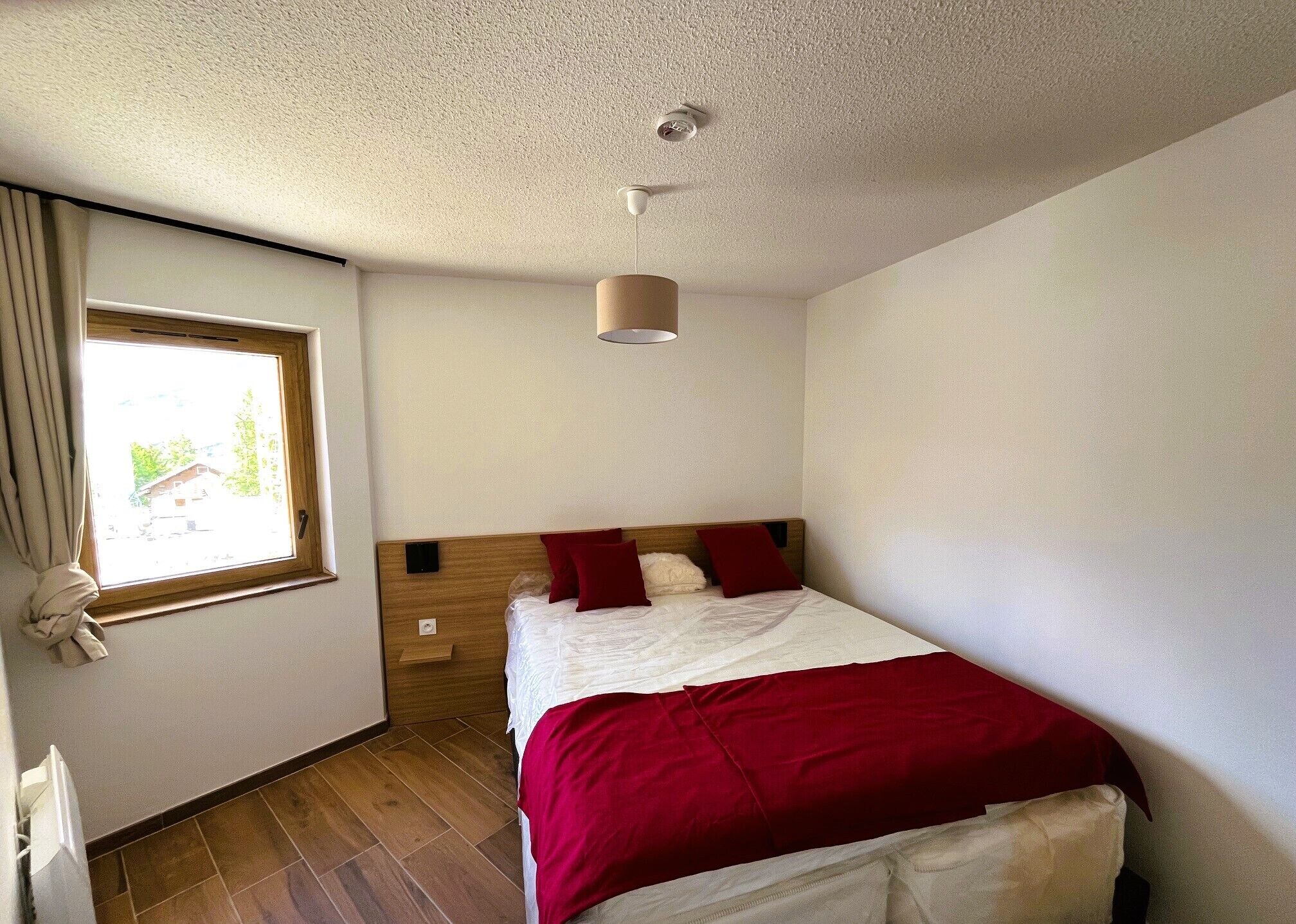 Une chambre à coucher avec une tête de lit en bois, un lit avec une literie rouge, une fenêtre avec des rideaux et un luminaire au plafond.