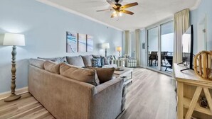 A modern living room with a sofa, coffee table, and lamps. There is a ceiling fan and a view of the outside through large windows. - Palacio 405 (Pensacola)