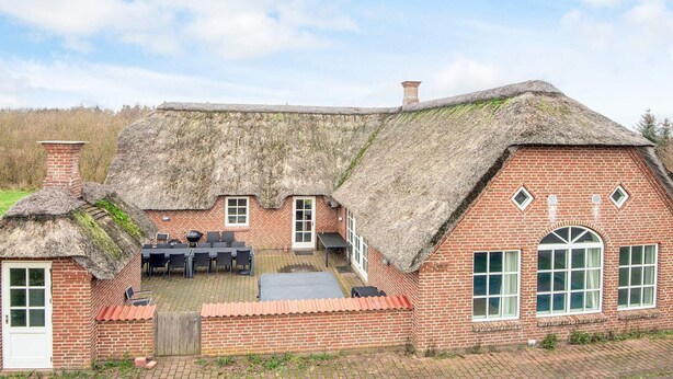 Une maison traditionnelle en briques avec un toit de chaume, une terrasse dotée de mobilier d’extérieur et une cheminée en briques.