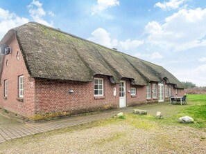 Une maison traditionnelle en briques, avec un toit de chaume, une allée pavée et un espace verdoyant agrémenté d’une table et de chaises.