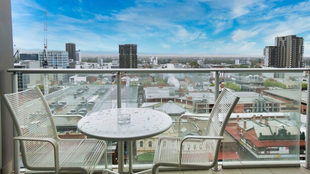 A balcony with a table and two chairs, overlooking a cityscape with various buildings and a clear sky. - Amyra Residences on Rowlands Place - Unit 1206 (Adelaide)