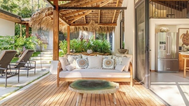 Eine Holzterrasse mit einem Reetdach und einer Sitzecke, eine Couch mit gemusterten Kissen sowie eine durch eine Glastür einsehbarer Küchenbereich.