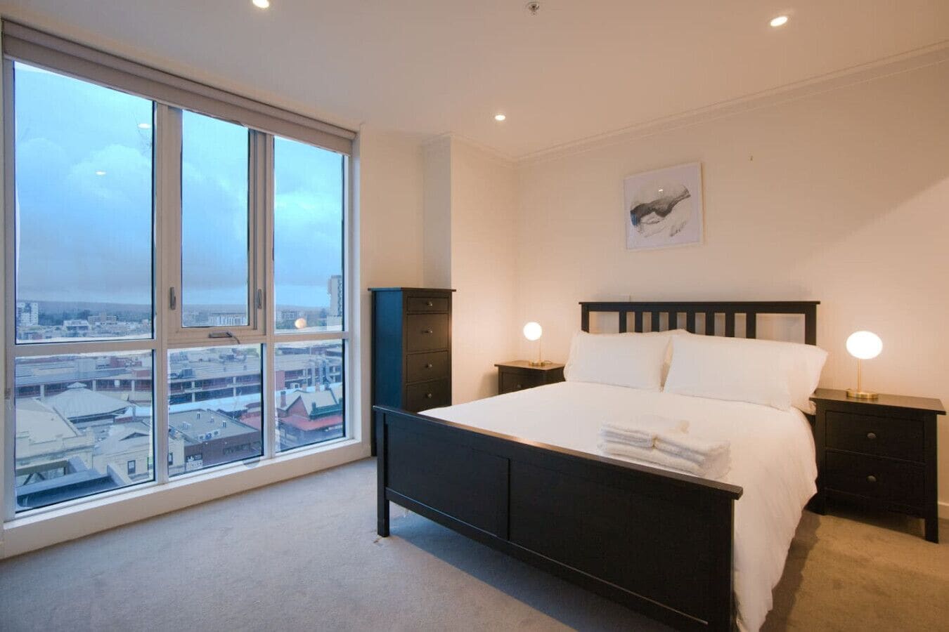 A bedroom with a large window, a bed, two bedside tables with lamps, and a city view.