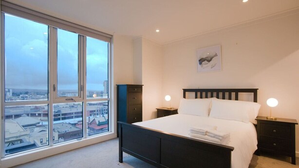 A bedroom with a large window, a bed, two bedside tables with lamps, and a city view. - Amyra Residences on Rowlands Place - Unit 906 (Adelaide)