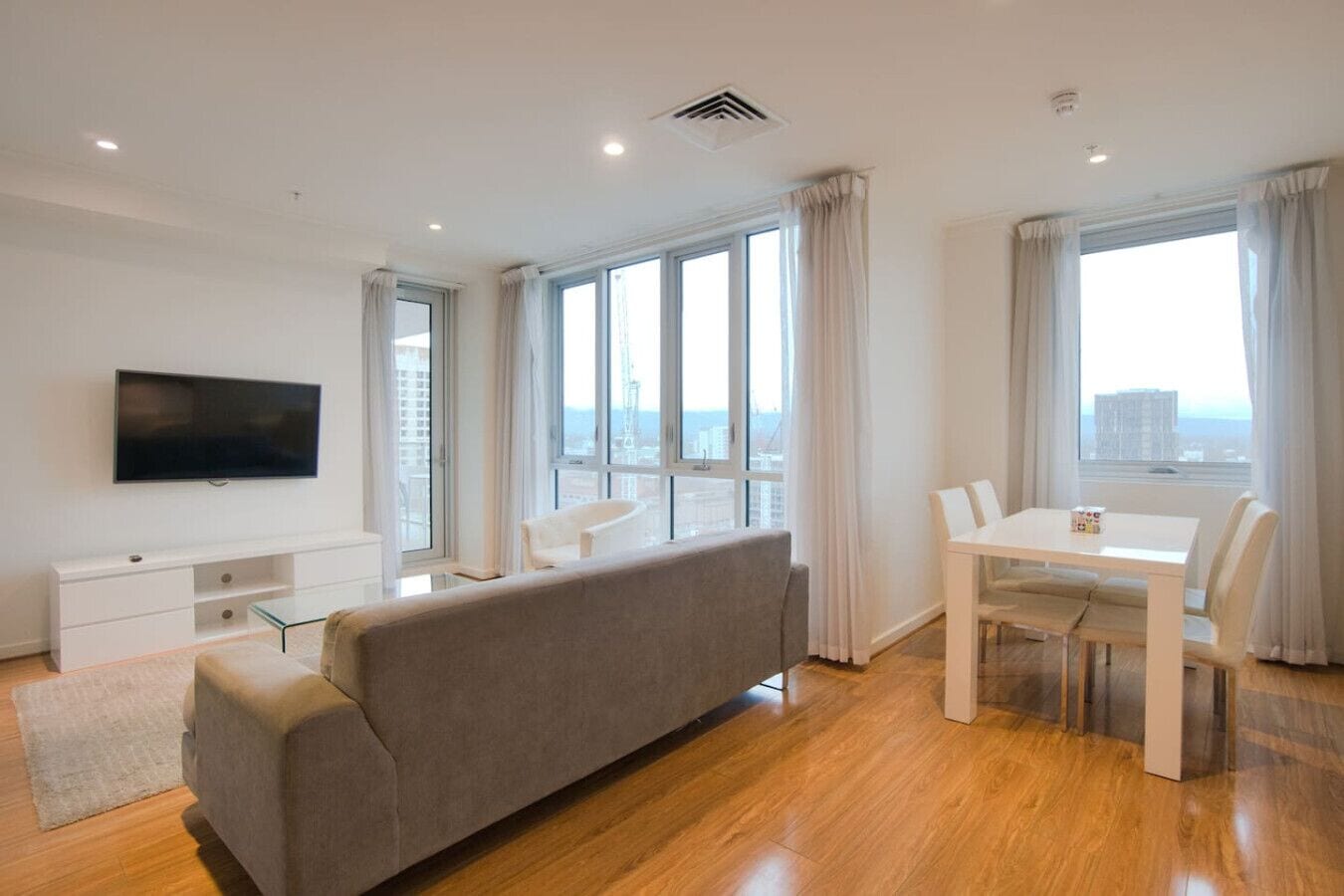 A modern living room with a sofa, TV, dining table, and large windows offering a city view.