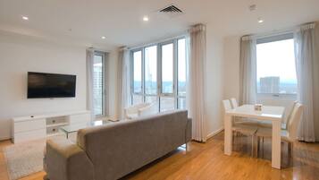 A modern living room with a sofa, TV, dining table, and large windows offering a city view.
