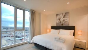 Ein Hotelzimmer mit einem großen Bett, zwei Nachttischlampen, Blick auf die Stadt durch raumhohe Fenster und einem Gemälde über dem Bett.