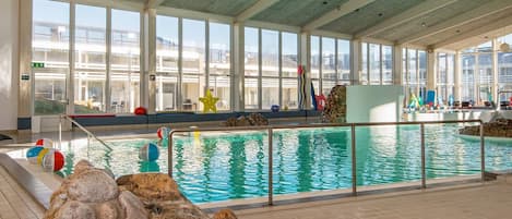 An indoor swimming pool with clear glass walls, a stone wall in the foreground, and a view of the outside buildings.