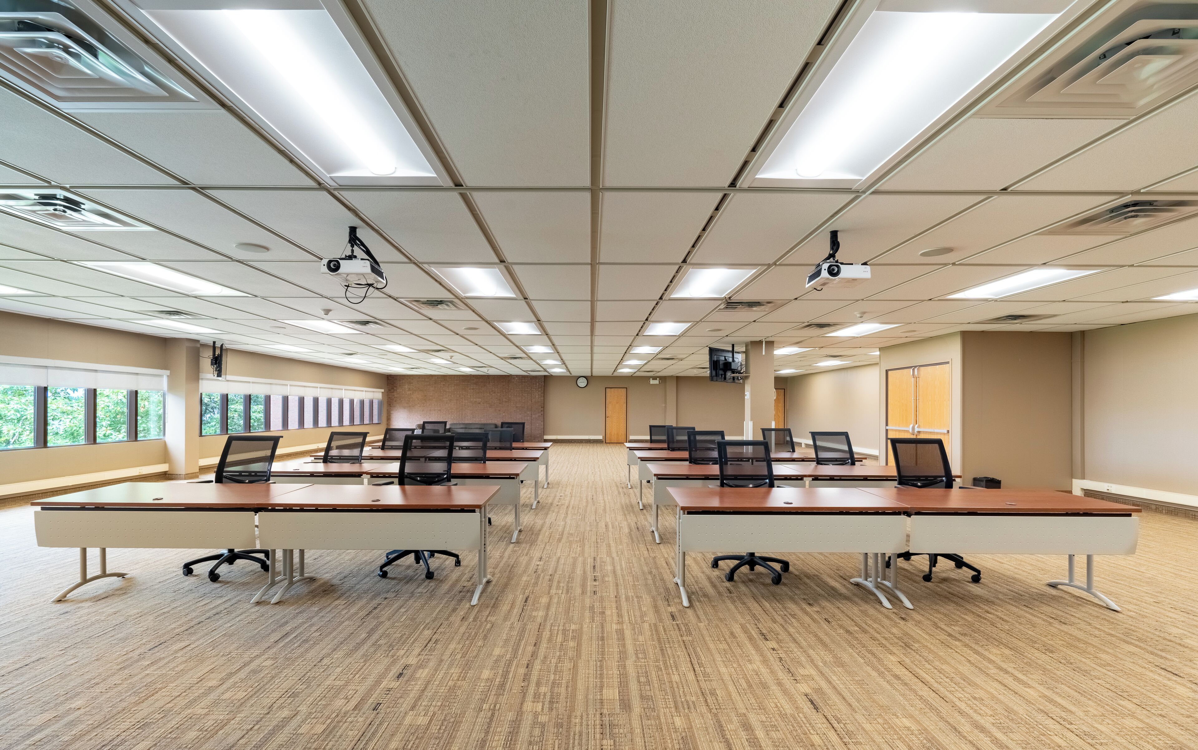 A conference room with multiple tables and chairs, ceiling-mounted projectors, and large windows with blinds.