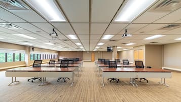 A conference room with multiple tables and chairs, ceiling-mounted projectors, and large windows with blinds.