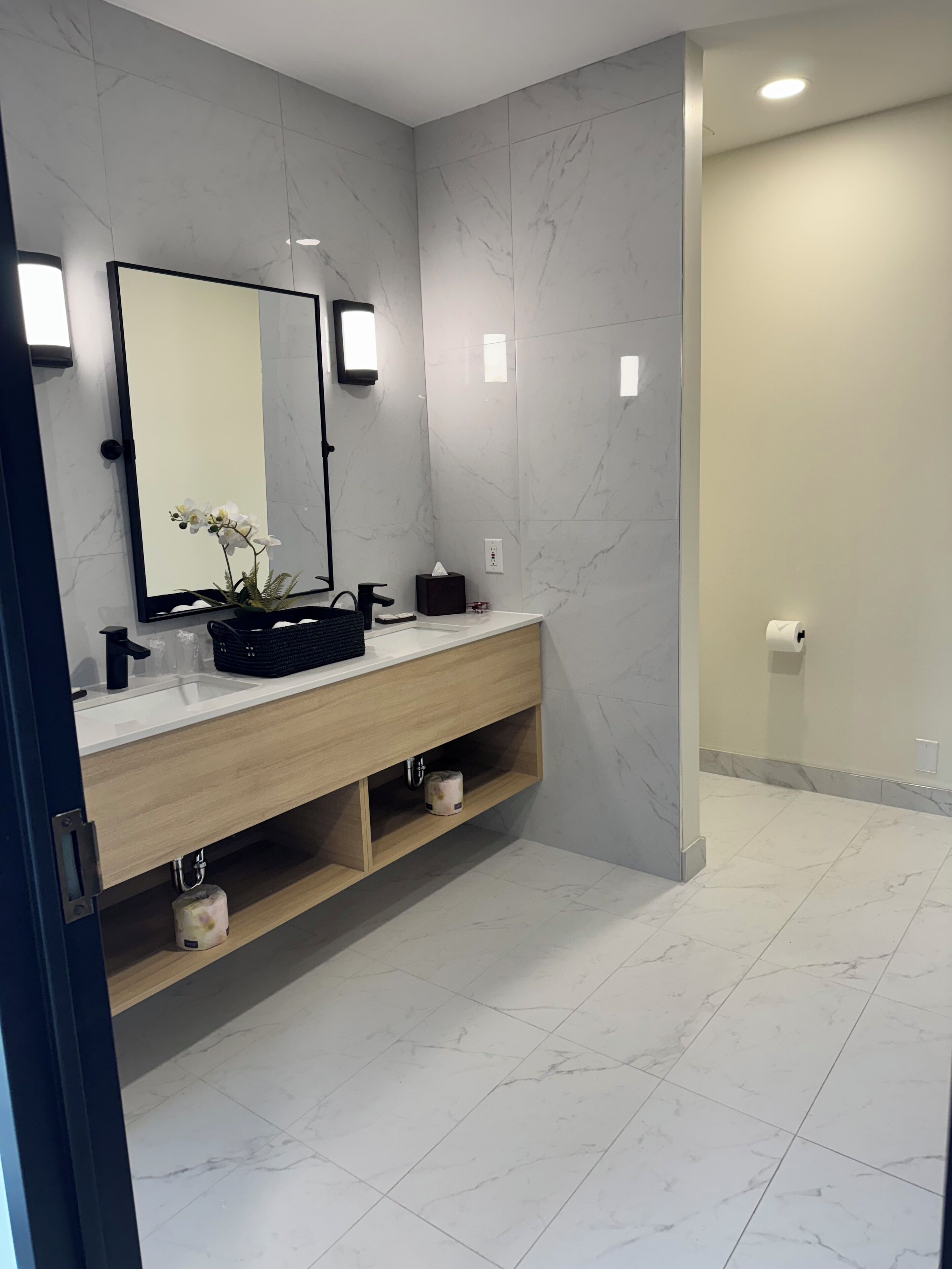 Une salle de bain moderne avec un mur recouvert de carreaux de marbre, un meuble-lavabo en bois, un miroir et une étagère ornée d’une plante.
