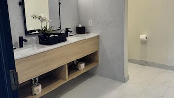 Une salle de bain moderne avec un mur recouvert de carreaux de marbre, un meuble-lavabo en bois, un miroir et une étagère ornée d’une plante.
