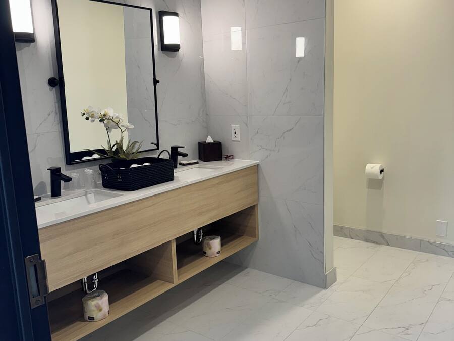 Une salle de bain moderne avec un mur recouvert de carreaux de marbre, un meuble-lavabo en bois, un miroir et une étagère ornée d’une plante.