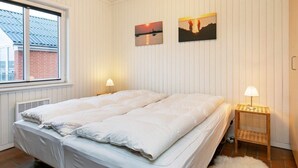 A double bed with white bedding, a window with blinds, two wall-mounted lamps, and two framed pictures.