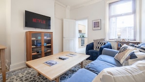 Ein Wohnzimmer mit Couchtisch aus Holz, einem blauen Sofa, einem Bücherregal und einem Fernseher, der 'NETFLIX' zeigt.