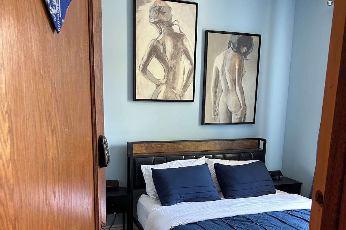 A bedroom with a wooden door, a bed with blue bedding, two framed artworks on the wall, and a small bedside table.