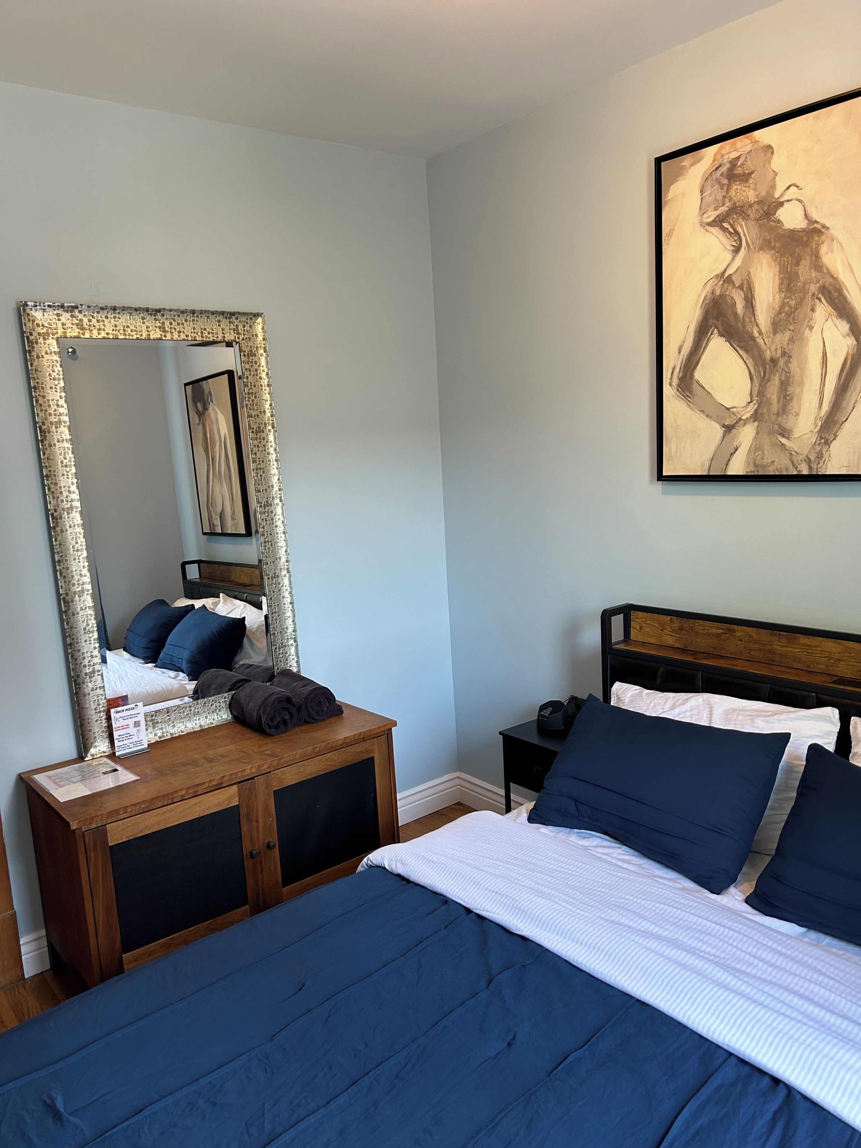 Un dormitorio con cama, mesita de noche, cómoda, espejo y un cuadro en la pared.
