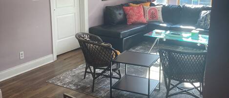 A living room with a black leather sofa, a glass coffee table, and a dining table with chairs. There is a ceiling fan, a framed picture on the wall, and a window with a view of buildings.