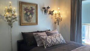 A bedroom with a bed, pillows, a painting, wall-mounted candelabras, and a window with curtains. - Dorothy's (Chicago)