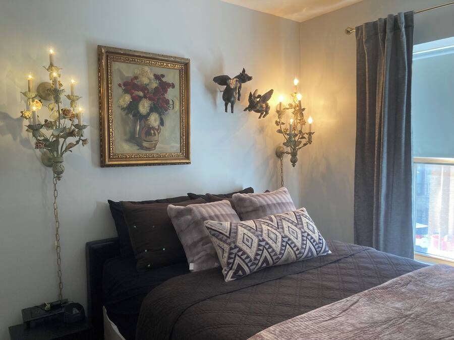 A bedroom with a bed, pillows, a painting, wall-mounted candelabras, and a window with curtains.