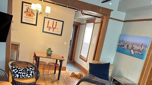 A living room with a black patterned rug, a glass coffee table, a wooden dining table with chairs, a small bench, and a cityscape painting on the wall. - Dorothy's (Chicago)