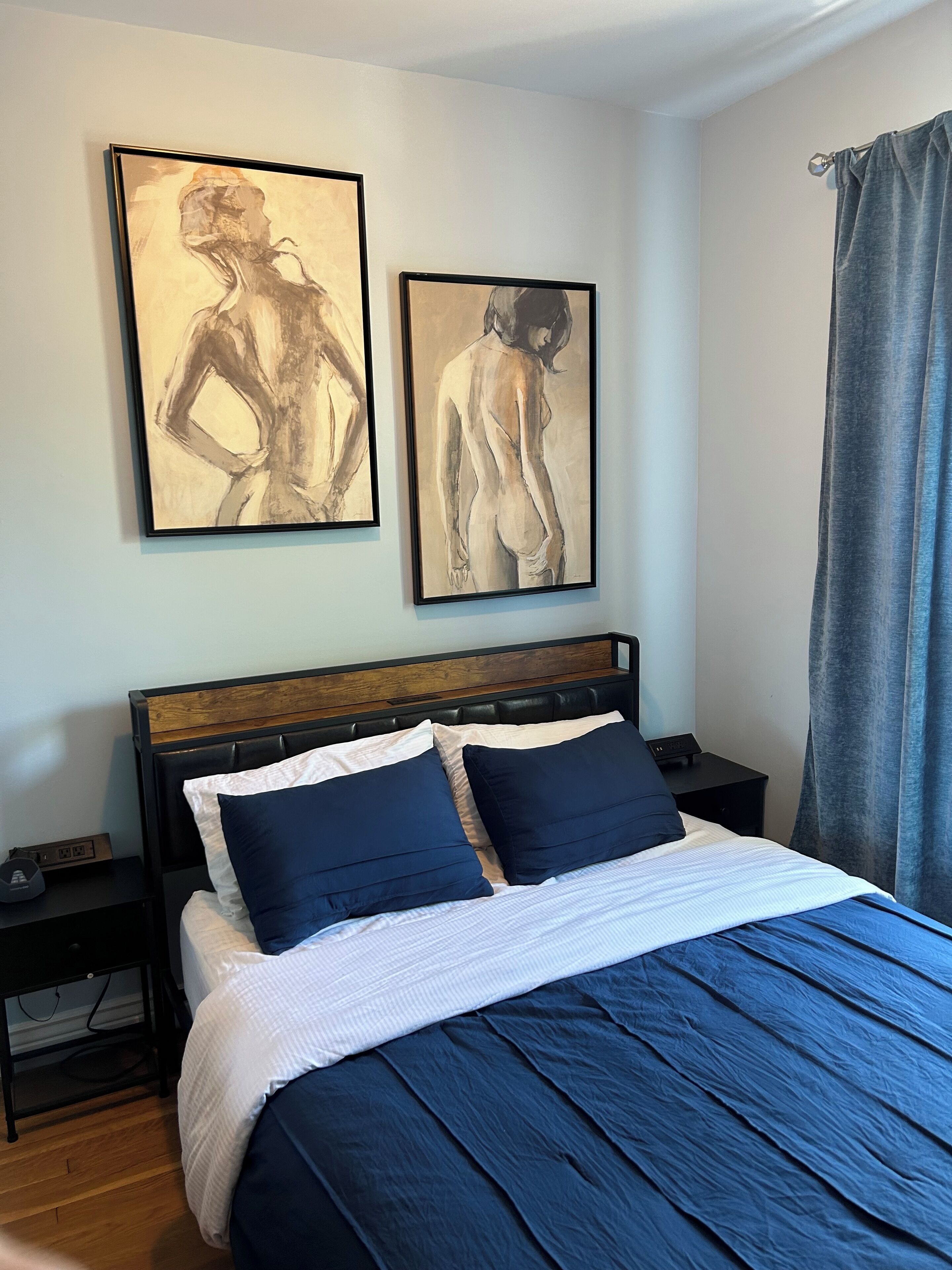 A bedroom with a bed, two nightstands, a wooden headboard, and two framed artworks on the wall.