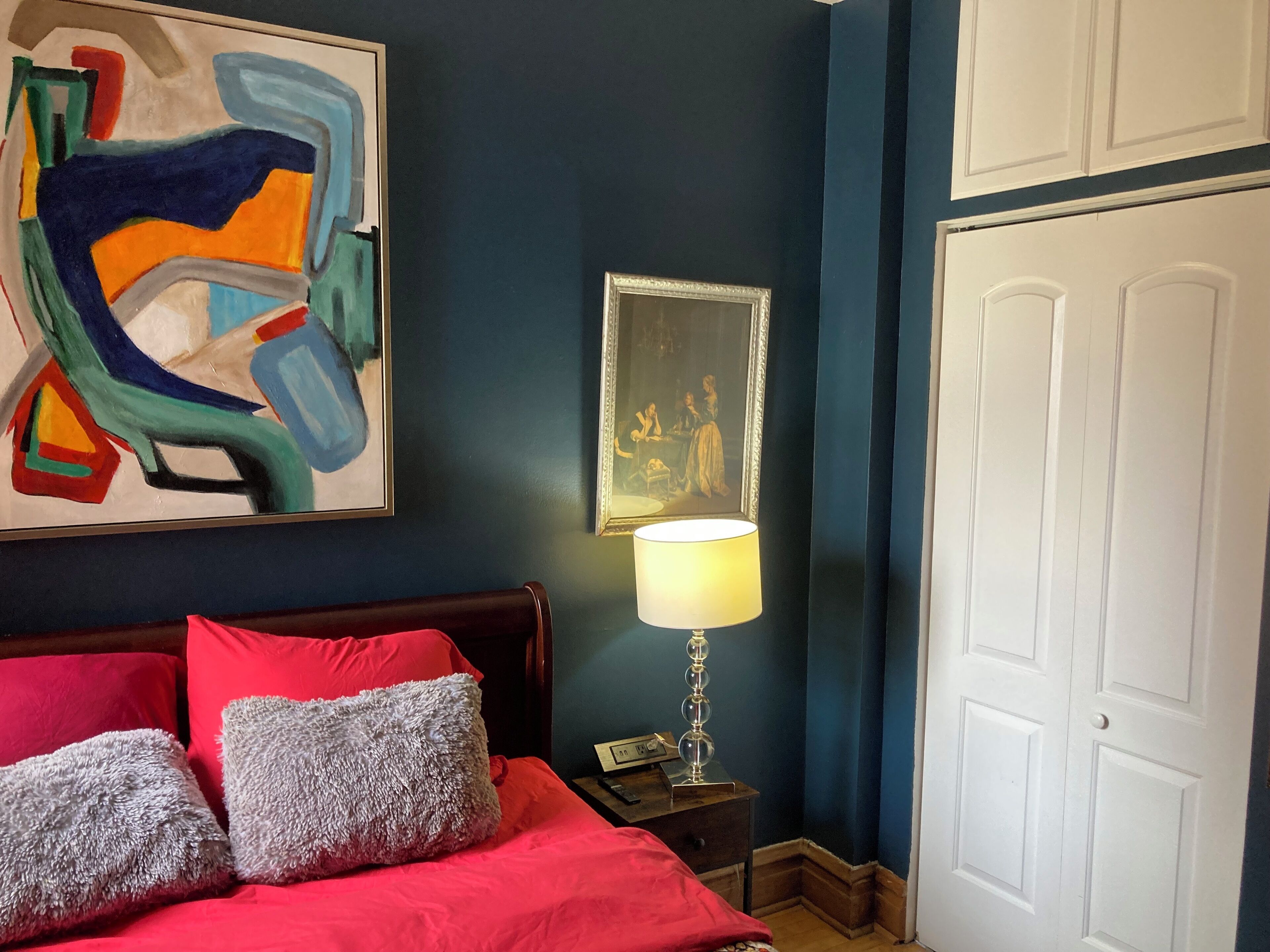 A bedroom with a bed, red and grey pillows, a painting, a lamp, and a framed picture on the wall.