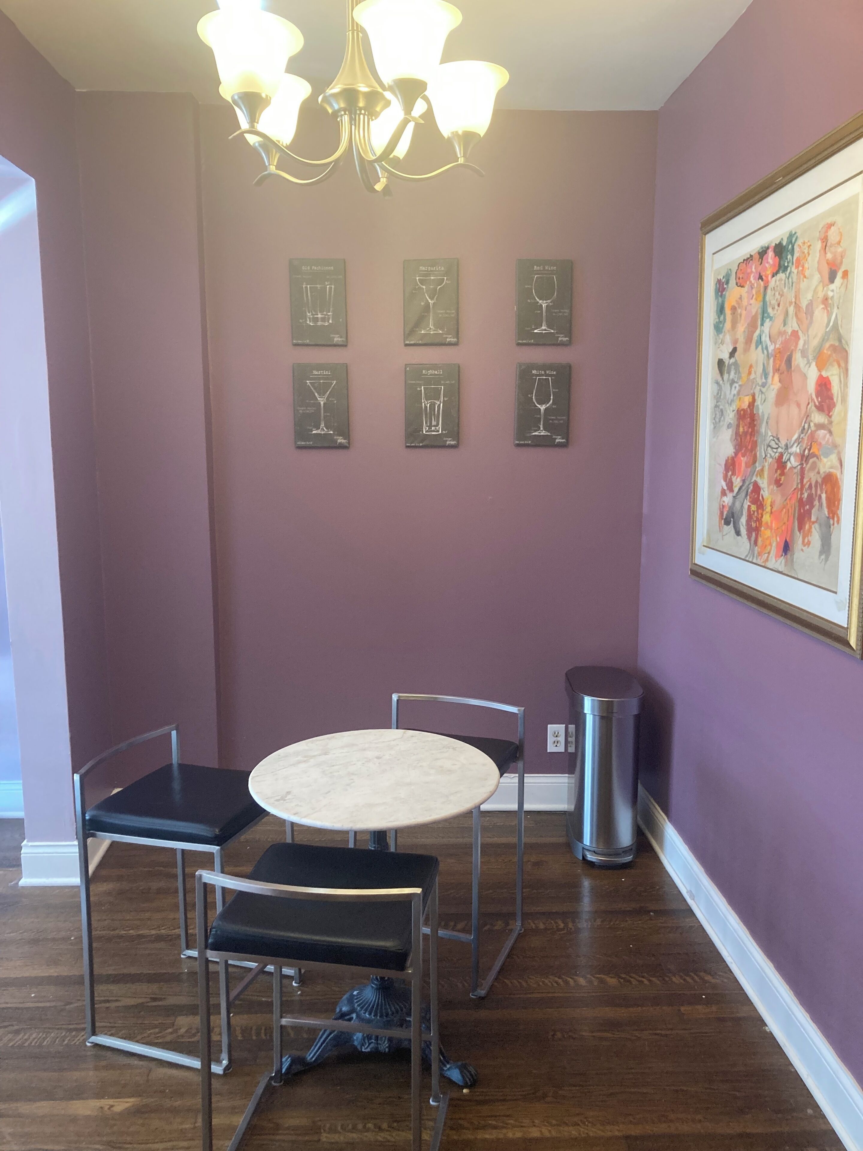 A dining area with a round table and four chairs, a chandelier, framed wine glass illustrations on the wall, and a trash can.