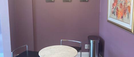 A dining area with a round table and four chairs, a chandelier, framed wine glass illustrations on the wall, and a trash can.