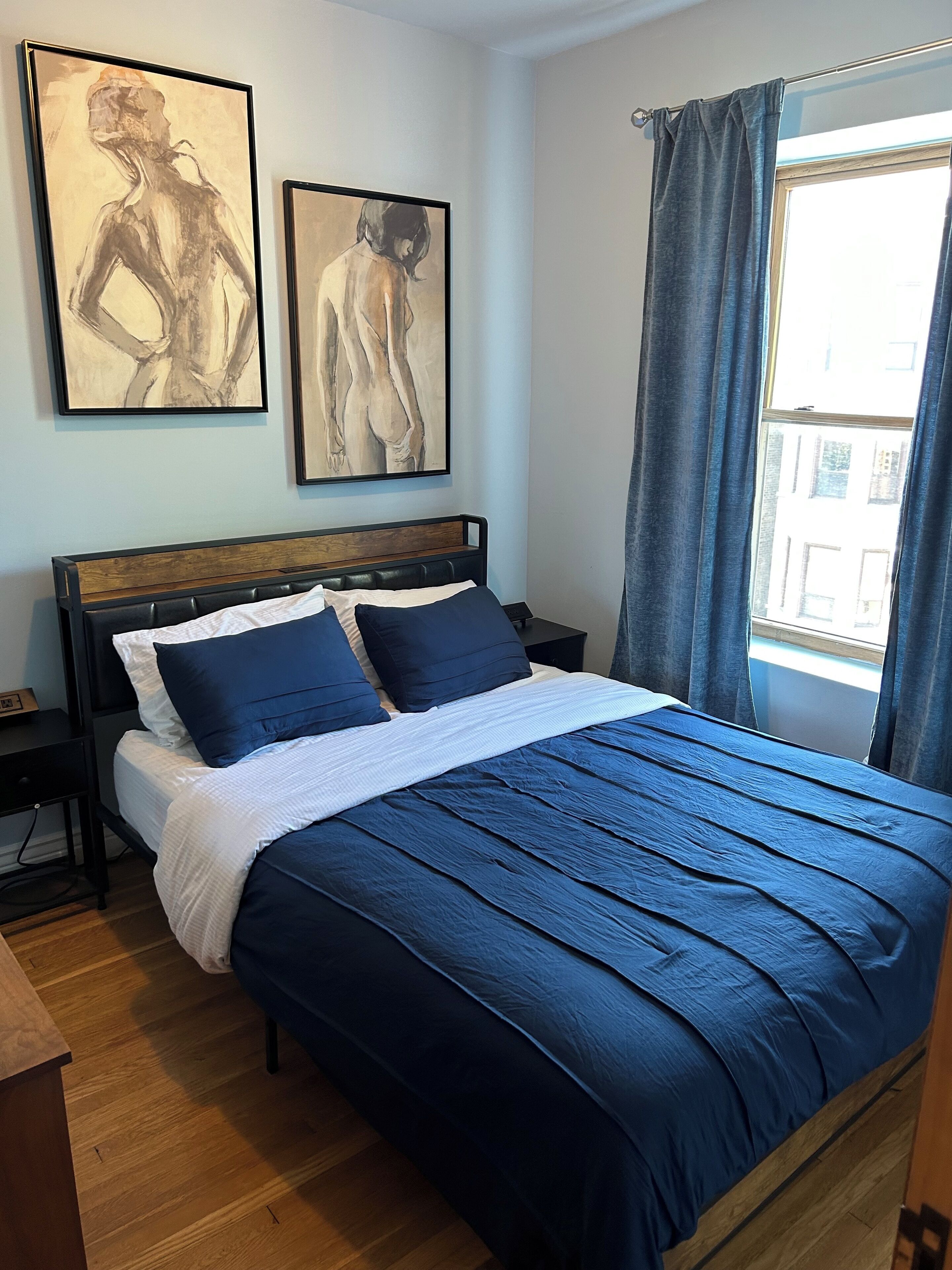 A bedroom with a large bed, two wall-mounted paintings, a nightstand, and a window with curtains.