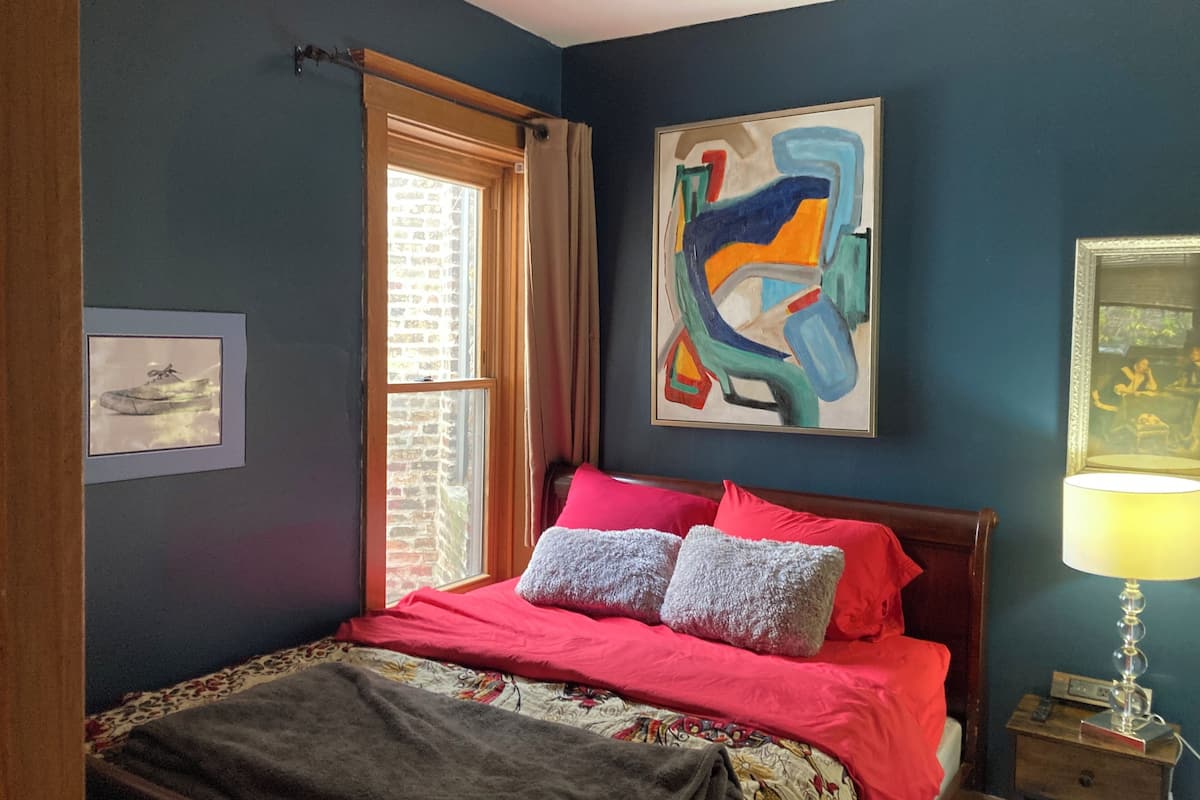 A bedroom with a bed, a window, a painting, and a nightstand.