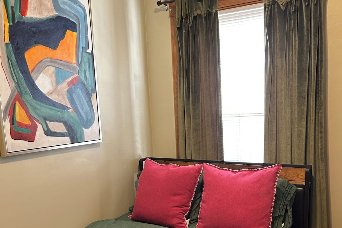 A bedroom with a bed, green and red pillows, a painting on the wall, and a window with curtains.