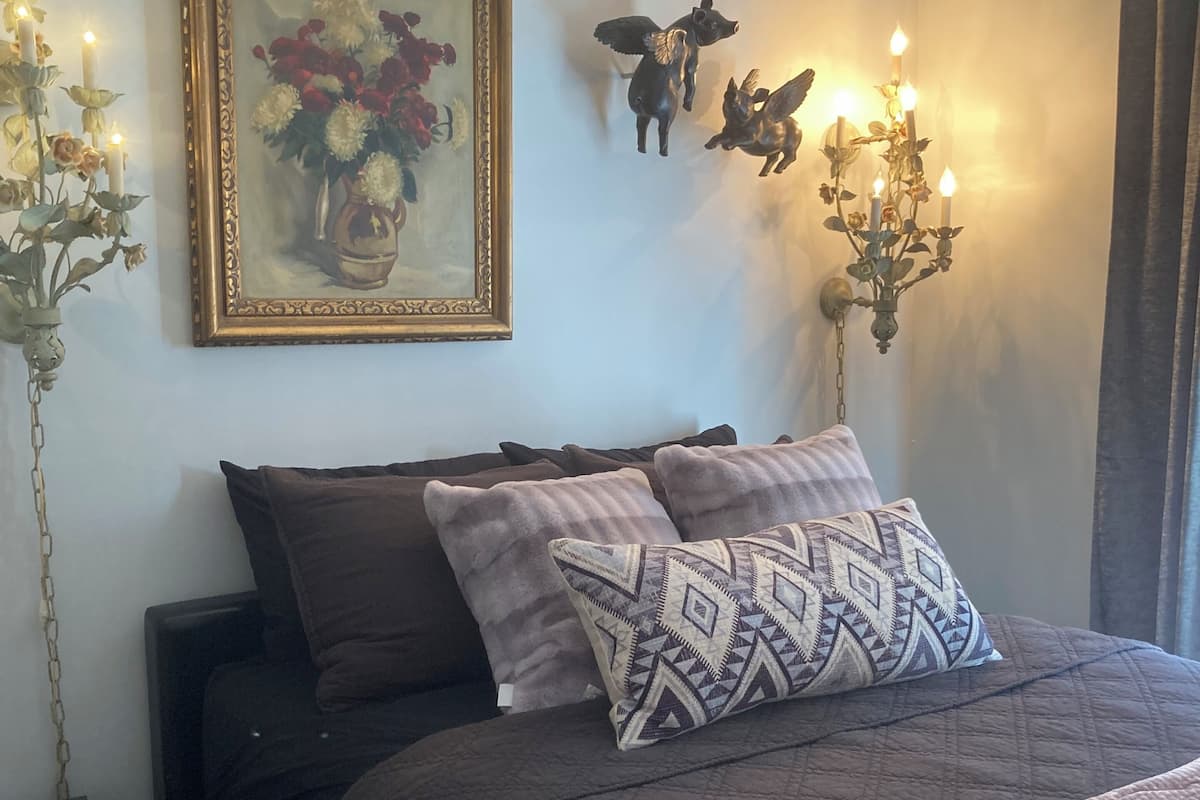 A bedroom with a bed, a nightstand, a lamp, a wall-mounted picture, and a wall sconce.