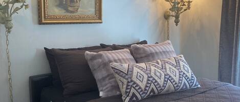A bedroom with a bed, a nightstand, a lamp, a wall-mounted picture, and a wall sconce.