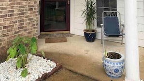 A covered porch with a brick wall, a sliding glass door, a potted plant, a blue chair, and a decorative urn.