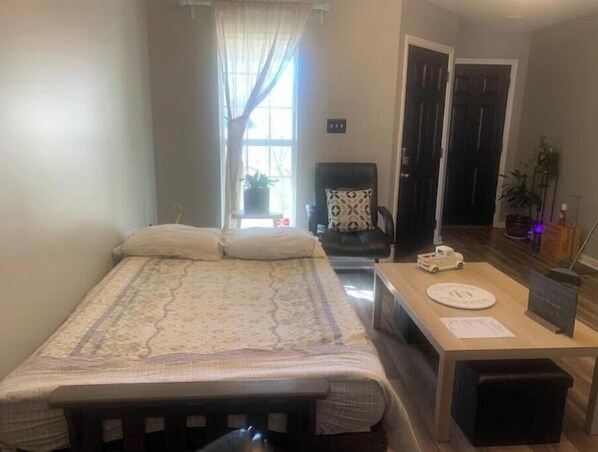 A small, well-lit room with a bed, a desk, and a chair. There is a window with curtains, a door, and a potted plant. - Green house (Southaven)