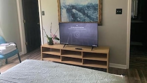 A hotel room with a bed, a television on a wooden stand, a framed picture on the wall, and a door to the left.