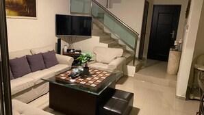 Ein modernes Wohnzimmer mit einem Glastisch, beigen Sofas, einem Flachbildfernseher und einer Treppe.