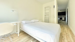 Un dormitorio moderno con una cama, un escritorio y una silla. Se ve un área de cocina al fondo.